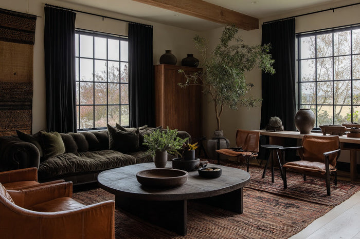 A stylish living room with a velvet sofa, leather armchairs, a round vintage coffee table, and large windows.