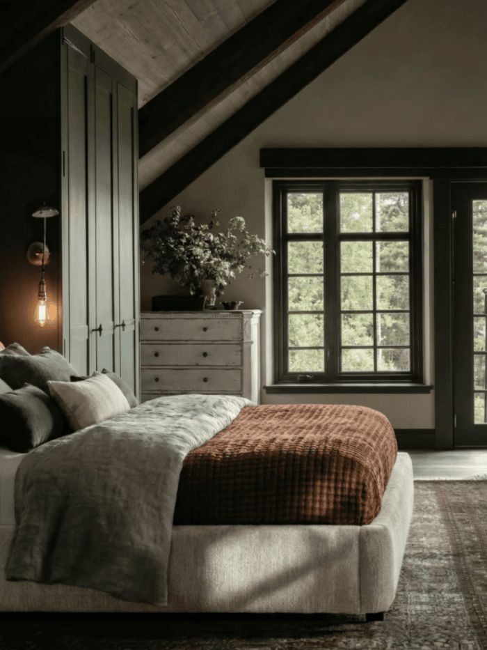 Moody modern rustic bedroom with upholstered bed, layered neutral bedding, plaid rust throw blanket, black-framed windows, and vaulted ceiling with exposed beams.