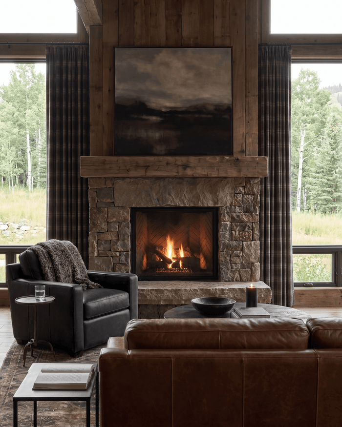 Modern rustic living room with a floor-to-ceiling stone fireplace, oversized landscape artwork, brown leather sofa, black leather lounge chair, round coffee table, and tall windows overlooking a wooded mountain landscape.