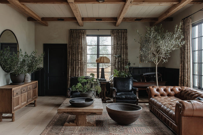Rustic living room with reclaimed wood coffee table, vintage wooden sideboard, leather sofa, black armchairs, large potted plants, and patterned rug.