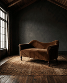 A vintage-style brown velvet chaise lounge with dark wooden legs, positioned on a distressed brown patterned area rug in a dimly lit attic room with dark textured walls and a window on the left. Modern Mountain living room custom furniture